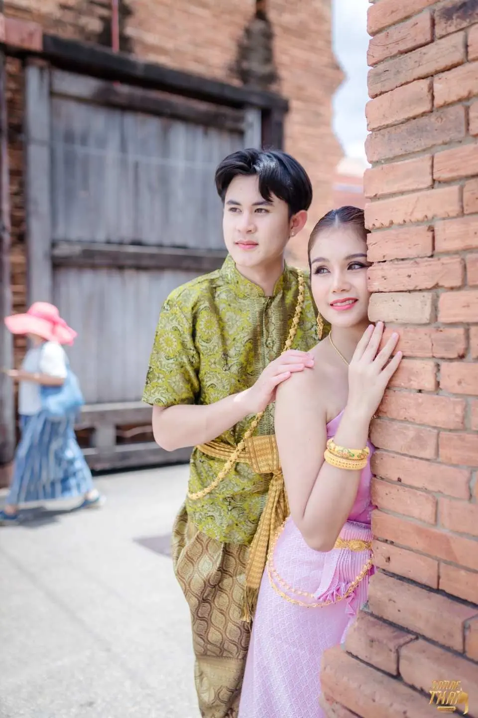 A couple in traditional Thai attire posing thoughtfully beside an ancient brick wall and wooden gate in Chiang Mai, creating a romantic historical scene.