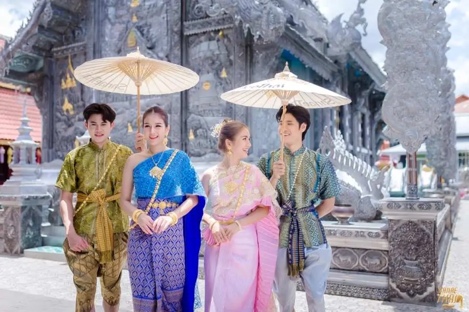 Four elegantly dressed visitors in traditional Thai garments holding classic umbrellas, posing in front of Chiang Mai's stunning Silver Temple (Wat Sri Suphan).
