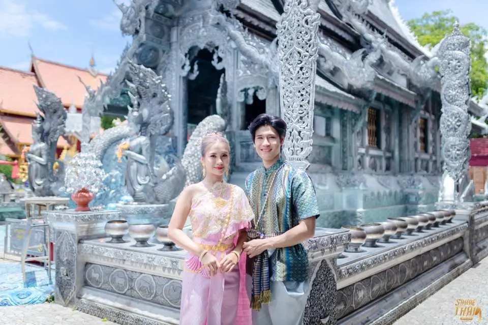 A happy couple in exquisite traditional Thai costumes standing proudly in front of the ornate Silver Temple (Wat Sri Suphan) in Chiang Mai, a perfect photo opportunity.