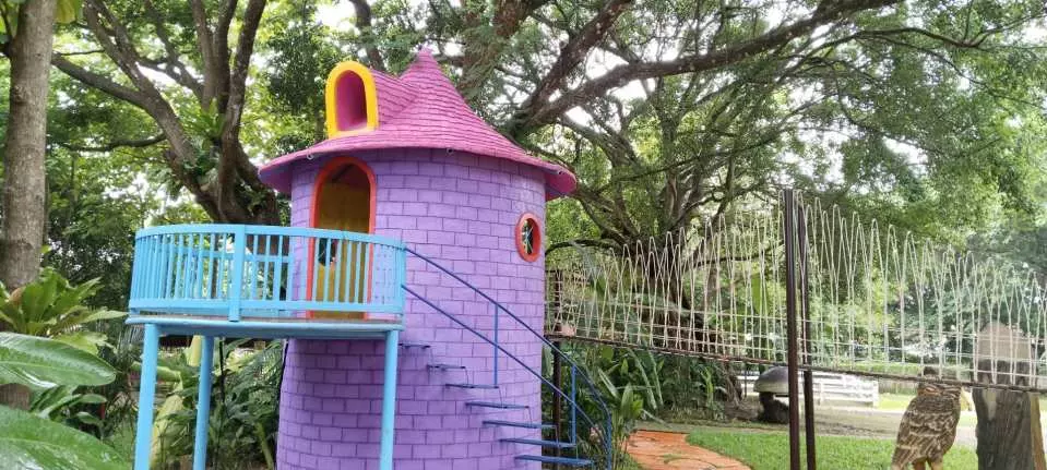 A whimsical purple castle-shaped children's playground with a pink roof and blue balcony staircase is nestled among lush green trees at Chiang Mai Hidden Village, a family-friendly attraction.