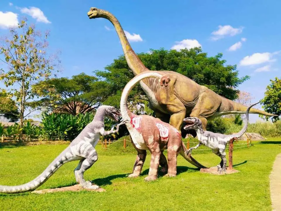 At Chiang Mai Hidden Village's dinosaur theme park, a tall Brachiosaurus sculpture stands with several smaller dinosaur sculptures on a vast green lawn, offering families fun exploration.