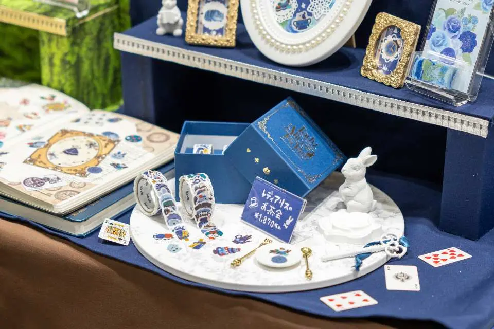 A whimsical Alice in Wonderland-themed stationery display at the Yokohama Stationery Girls Expo, featuring intricate washi tapes, decorative items, and a white rabbit figurine in blue and white tones, exuding playful charm.