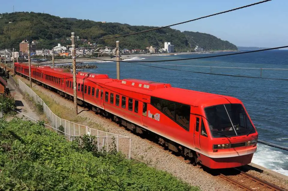 A vibrant red Resort21 sightseeing train travels along the coastal railway of Japan's Izu Peninsula, with the deep blue sea on the right and lush green hills on the left, creating a picturesque scene.