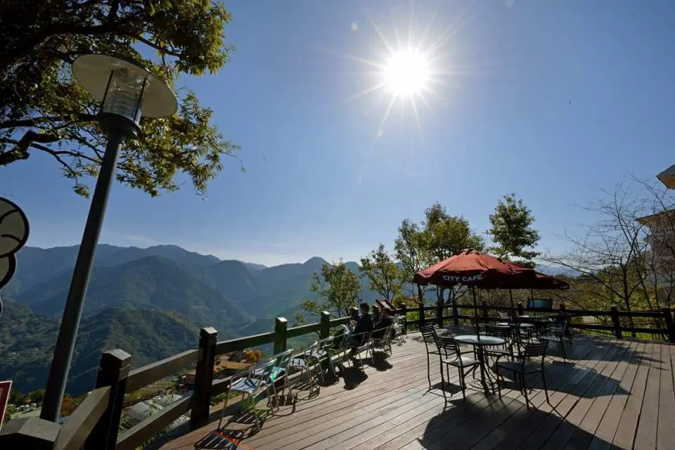 An outdoor wooden viewing deck at Cingjing Farm in Nantou, Taiwan, with tables, chairs, and an umbrella, offering spectacular mountain views under bright sunshine, perfect for enjoying coffee and scenery.