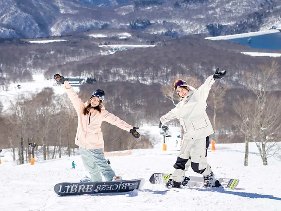Two happy snowboarders pose on the vast snowy slopes of Tambara Ski Park, with majestic snow-capped mountains and a lake in the background, fully enjoying the snowboarding experience.