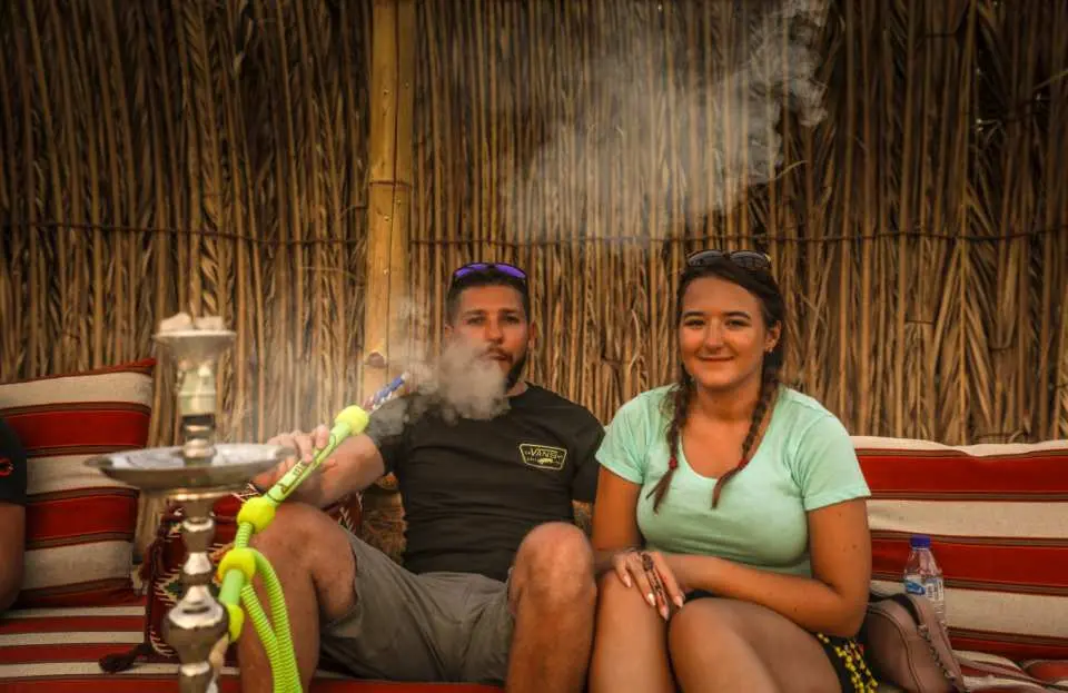 Tourists relaxing and enjoying traditional shisha in a Bedouin tent at an Abu Dhabi desert camp, experiencing the leisurely local customs.