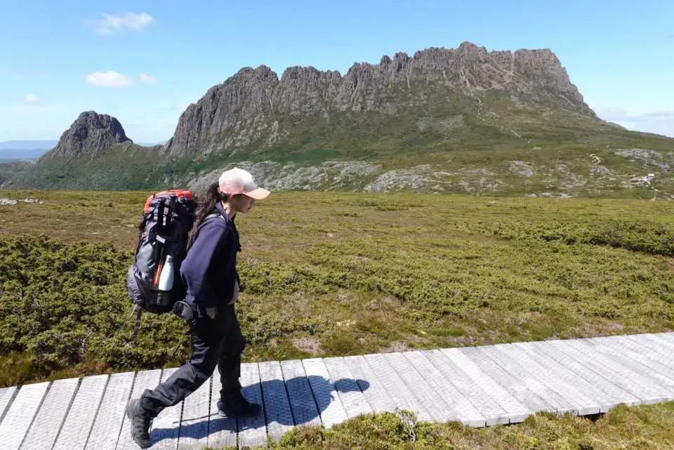 Hiker with a large backpack on a wooden boardwalk, exploring the stunning wilderness of Cradle Mountain National Park, with rugged peaks dominating the Tasmanian landscape.