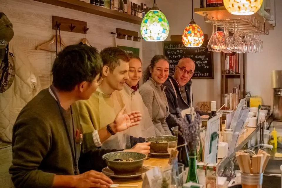 During a private guided Sapporo ramen culture tour, the guide interacts with visitors at a ramen shop counter, delving into ramen's history and culture, enjoying the fusion of knowledge and gastronomy.