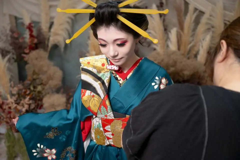 東京近郊の花魁変身体験で、女性がスタイリング過程に集中し、青緑色の着物と独特の髪飾りを身につけ、美しい花魁へと変身する瞬間を写真家が捉えています。