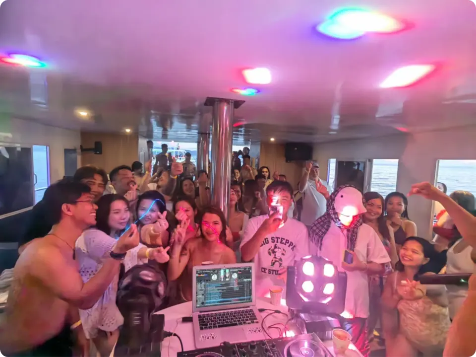 Young, enthusiastic guests dancing and celebrating on the Boracay yacht's interior deck at night, enjoying live DJ music under colorful party lights.