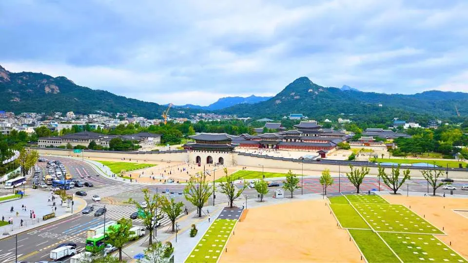 A panoramic elevated view of Gyeongbokgung Palace in Seoul, showcasing its grand architecture, surrounding city landscape, and distant mountains, as seen from a secret observatory.