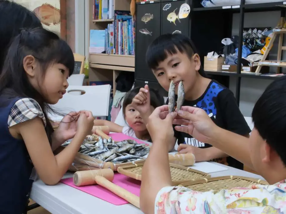 澎湖敲魚乾親子團體活動，小朋友們圍繞桌邊，好奇地觀察和學習如何處理魚乾，展現歡樂互動與共同探索傳統技藝。