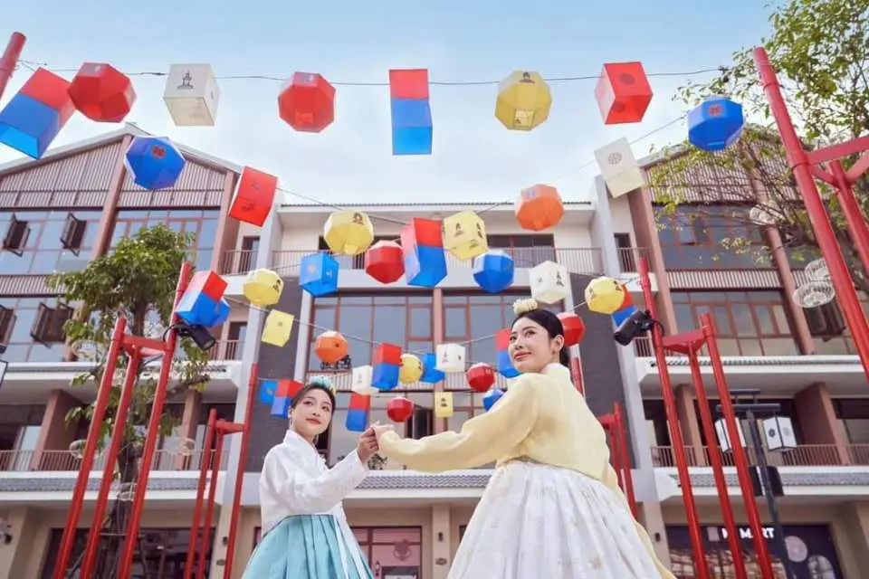 Two women in Hanbok enjoy a cultural experience in Hanoi Grand World's Korean-style district, surrounded by traditional lanterns hanging overhead.