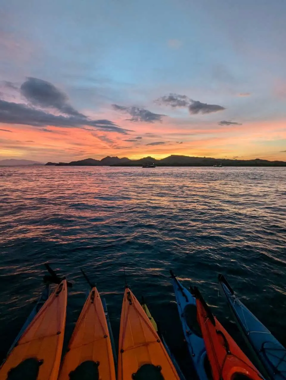 Several colorful kayaks are parked on the water in Labuan Bajo, facing a breathtaking sunset over Komodo National Park, concluding a perfect day of adventure.