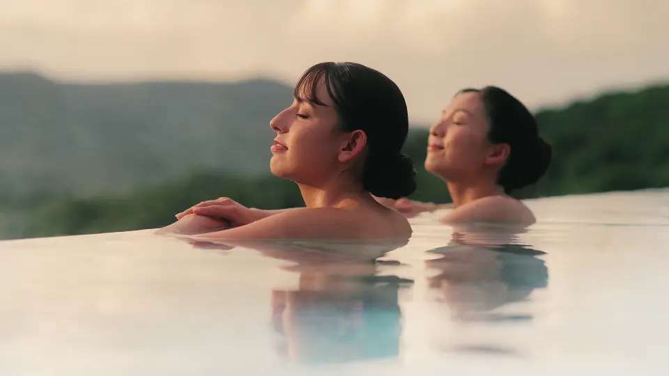 오키나와 정글리아 인피니티 온천에서 두 여성이 따뜻한 온천수와 노을을 즐기며, 울창한 산들을 배경으로 평화롭고 낭만적인 휴식의 순간을 만끽하는 모습.
