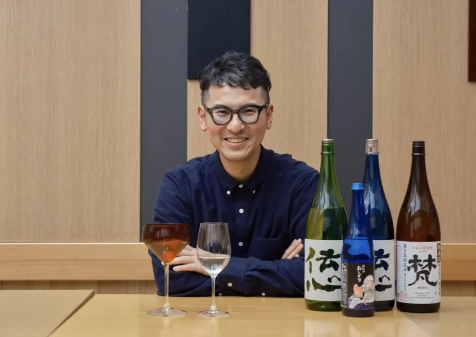 The friendly owner of Kubota Saketen in Sabae, Fukui, posing with local sake bottles and unique lacquerware cups, ready to guide guests through their tasting journey.
