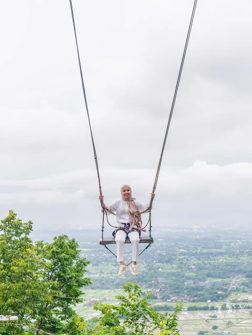 Visitor enjoying the thrill on a giant swing at Obelix Hills, smiling with a panoramic view of Yogyakarta's lush green mountains in Indonesia.