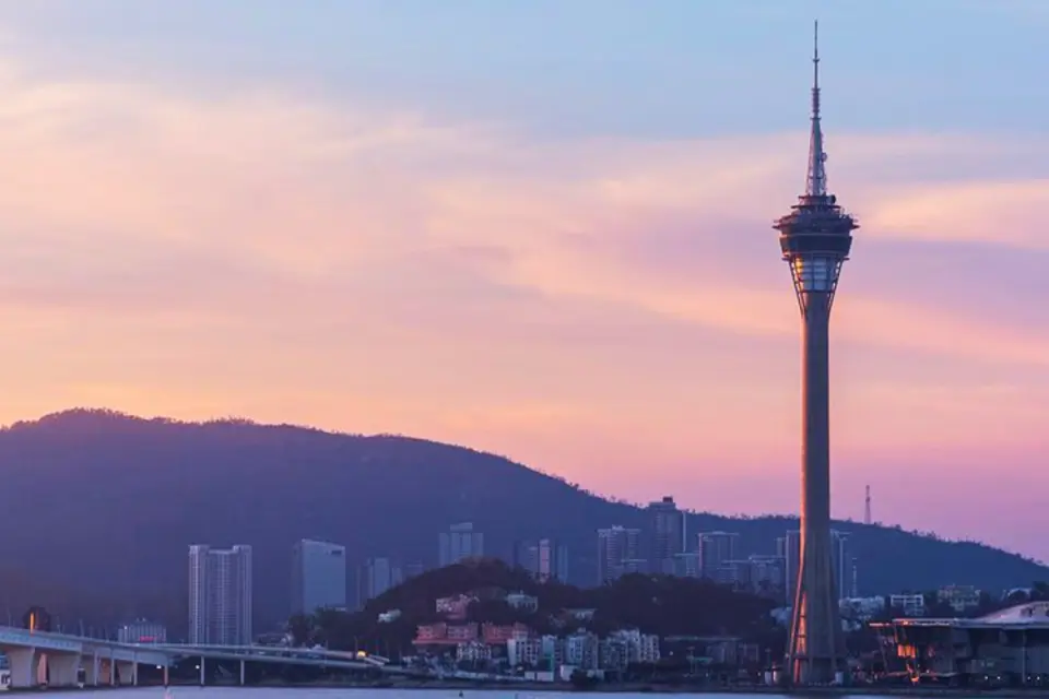 Macau Tower against a pink and purple sunset sky, adorned with surrounding mountains and buildings, an iconic sightseeing landmark in Macau.