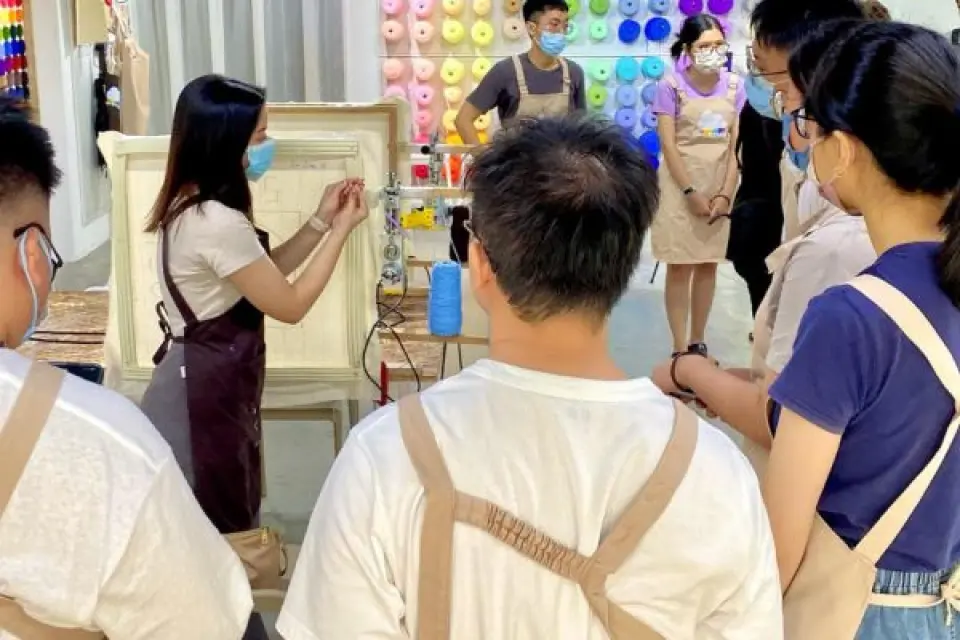 槟城OLO Studio簇绒工作坊中，导师向戴口罩的学员示范簇绒技巧，背景有彩色纱线墙，呈现新手友好的实作体验。