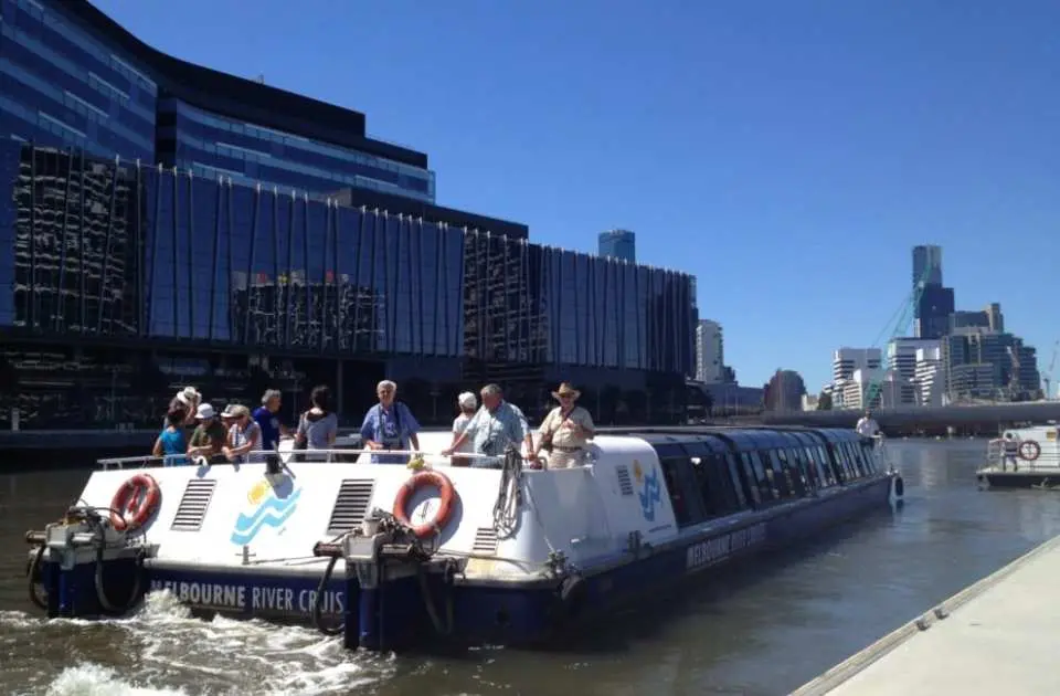 雅拉河观光游船上的乘客在露天甲板享受阳光，近距离欣赏墨尔本现代玻璃幕墙建筑与城市风光。