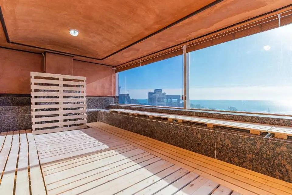 Busan Haeundae HILL SPA's modern sea view jjimjilbang, featuring wooden floors and a relaxation area, where guests can enjoy unique sweating experience while admiring Haeundae's blue ocean through large windows.