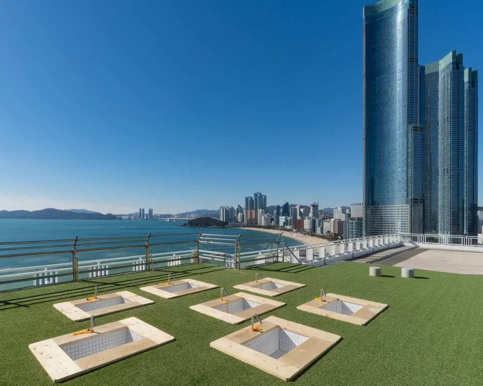 Busan Haeundae HILL SPA's rooftop outdoor relaxation area, featuring green artificial turf with square seating spaces, overlooking the vast ocean and Haeundae city skyline.