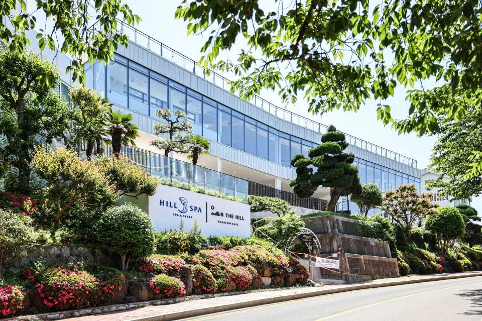 Busan Haeundae HILL SPA's lush green building exterior, surrounded by blooming flowers and well-manicured trees, showcasing its unique charm of combining spa services with natural landscapes.
