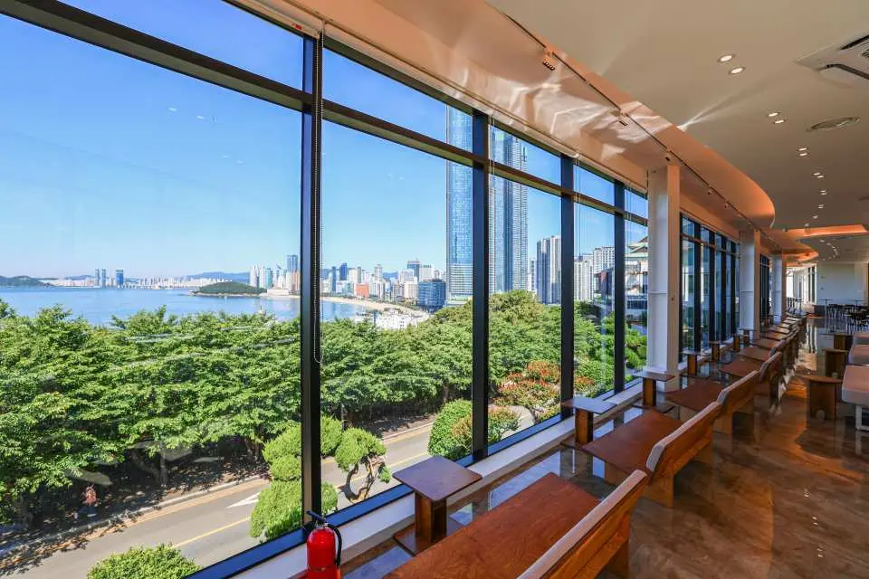 Scenic dining area inside Busan Haeundae HILL SPA, with comfortable window-side seating overlooking lush trees, busy streets, and the distant Haeundae sea view and city skyline.