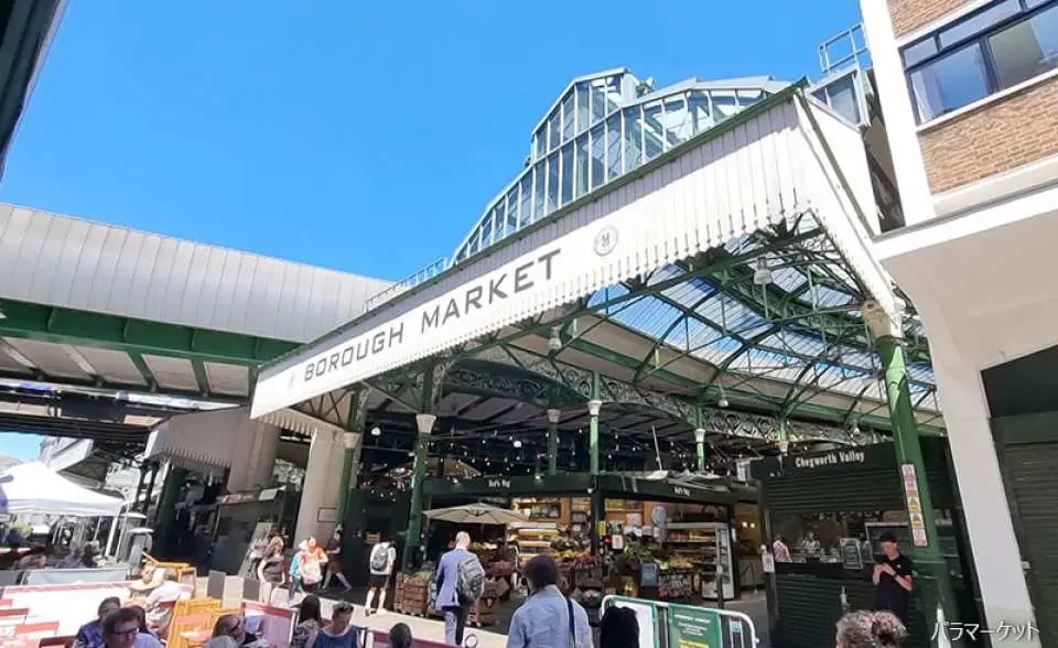 ロンドンの有名なグルメ市場、バラ・マーケットのにぎやかな入り口と特徴的な建築。アシスタントとともに多様な地元産品を探索する訪問者。