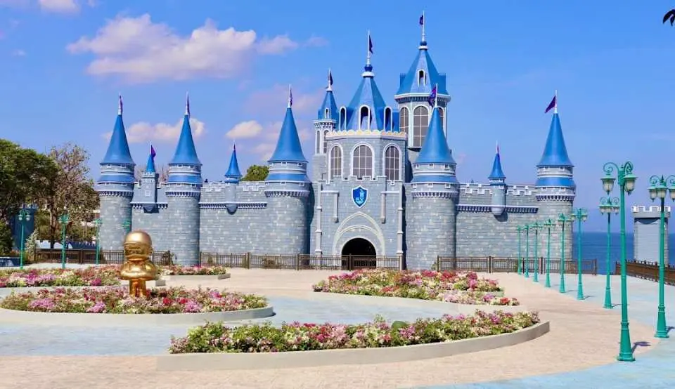 The majestic blue and grey JCastles theme park exterior in Batangas, Philippines, featuring enchanting turrets, lush flowerbeds, and a golden fountain, welcoming families to adventure.