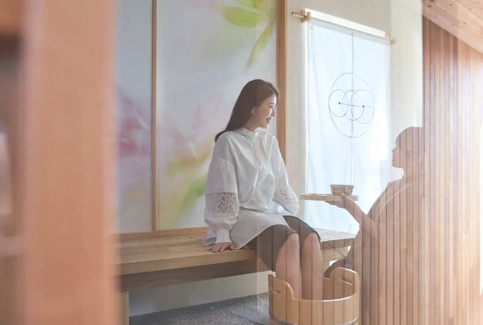 A happy guest enjoying a relaxing foot bath experience at Arashiyu Kamakura, receiving warm service and tea from a therapist in a sunlit, traditional setting.