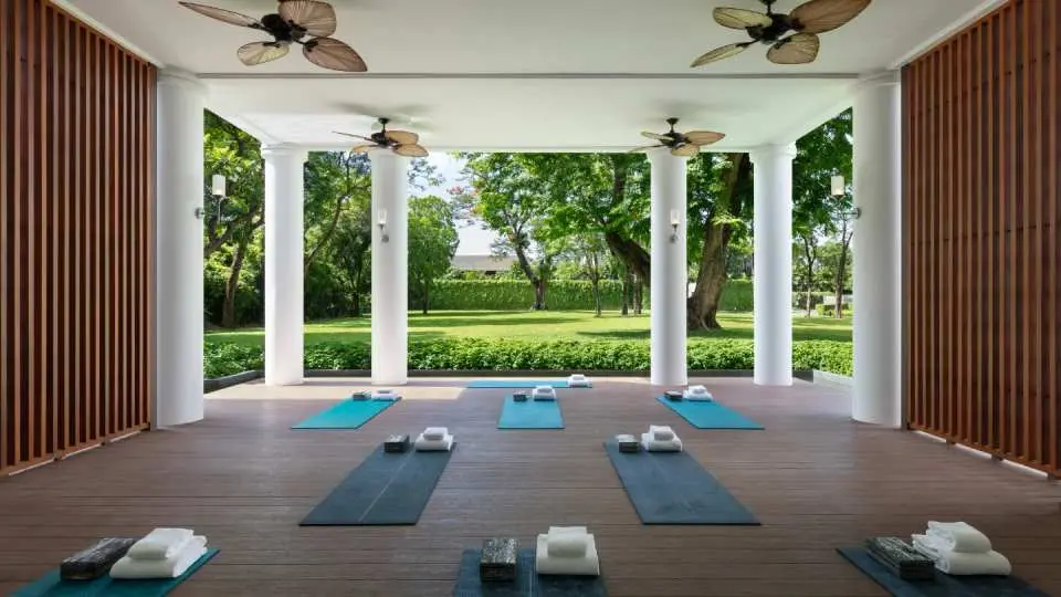 Open-air yoga pavilion at The Sukhothai Bangkok's spa, with multiple yoga mats on a wooden floor, inviting guests to relax and rejuvenate amidst lush garden views.