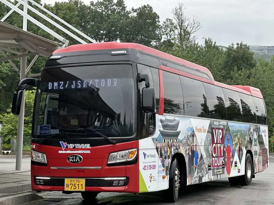 紅白色VIP城市之旅巴士，車身標示「DMZ/JSA TOUR」，象徵從首爾出發前往DMZ地區的便捷交通。
