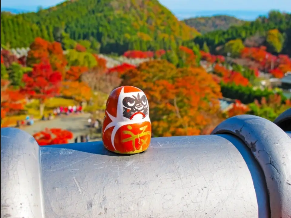 勝尾寺の勝利だるまが手すりの上に座り、背景には秋限定の鮮やかな紅葉が広がり、遠くの谷の景色を眺めます。