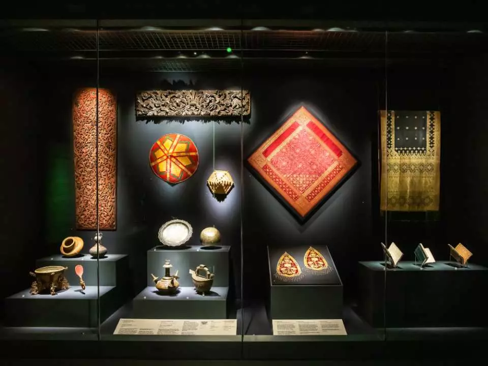 Inside the Peranakan Museum Singapore, a display case exhibits various Peranakan handicrafts, including lanterns, textiles, pottery, and gold ornaments.