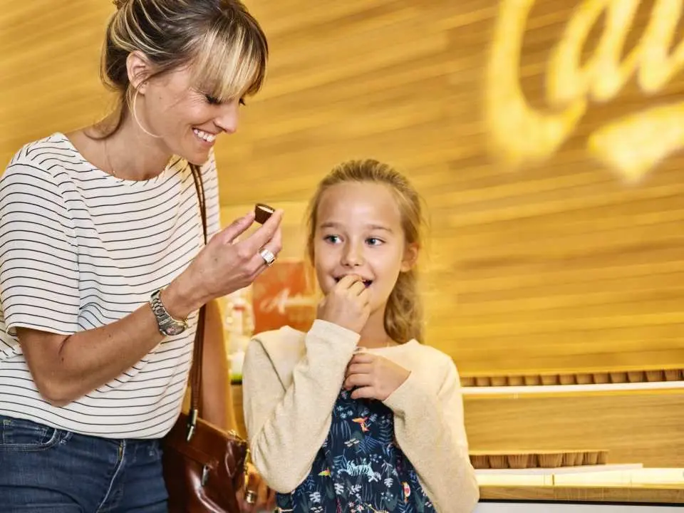 A mother and daughter enjoying a sweet tasting experience at Maison Cailler chocolate factory, with the mother offering chocolate and the daughter's face full of anticipation and joy.