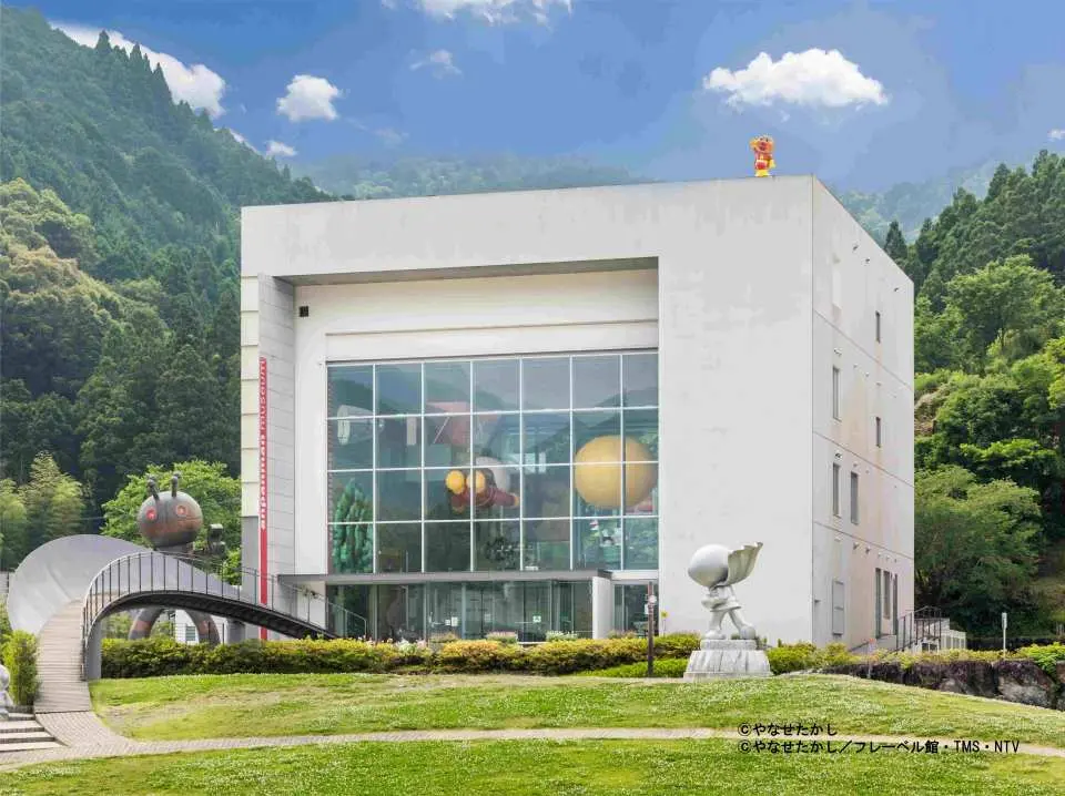 Exterior view of the modern Takashi Yanase Memorial Museum in Kami City, Japan, featuring large glass windows, unique sculptures, and lush green mountains in the background, a key stop on the Yanase world tour.
