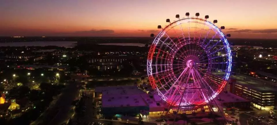 The iconic Orlando Eye Ferris wheel, brilliantly illuminated with patriotic red, white, and blue lights, stands majestically against a stunning sunset over the city skyline.