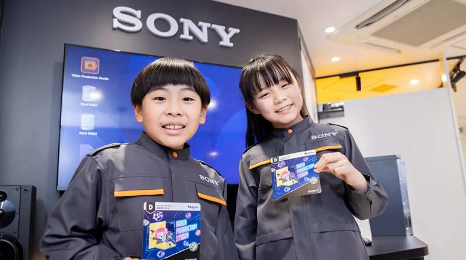 キッザニア甲子園のSONY施設で、ブランド制服を着た2人の子供たちが製品設計図を持ち、テクノロジー製品の開発とデザインの仕事を体験し、イノベーションを学んでいます。