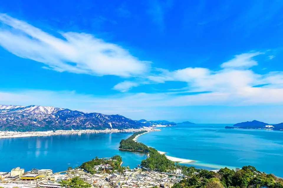 A breathtaking panoramic view of Amanohashidate from the observation deck, one of Japan's Three Scenic Views, showing the pine-covered sandbar across Miyazu Bay with distant snow-capped mountains.