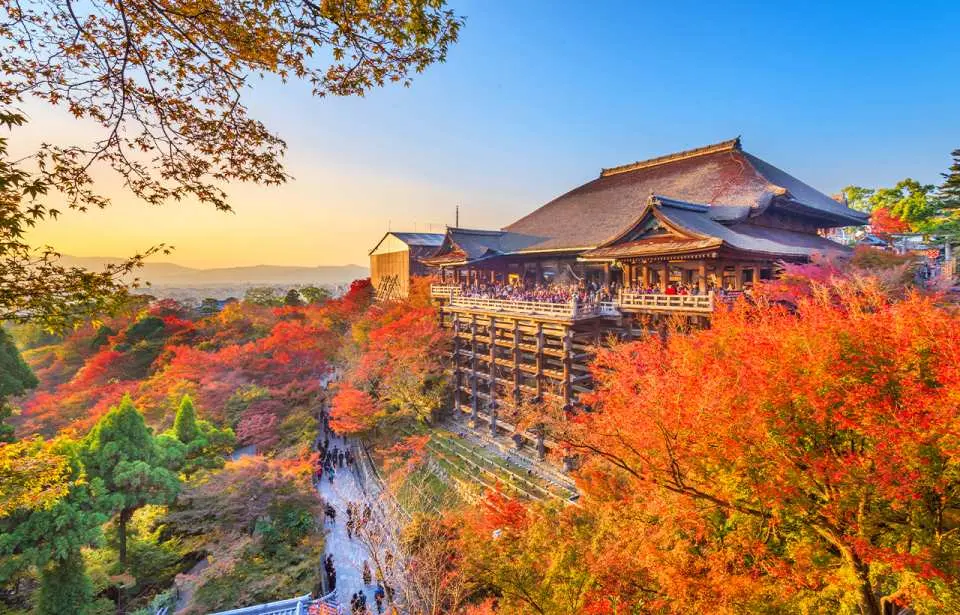 The iconic Kiyomizu-dera Temple in Kyoto, with its wooden scaffolding, surrounded by vibrant red and orange autumn foliage under a beautiful sunset sky, showcasing traditional Japanese architecture.