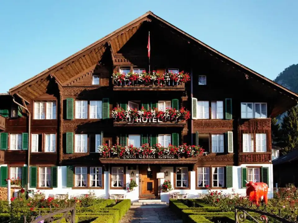 Exterior of a traditional Swiss chalet-style hotel, adorned with flower-filled balconies and green shutters, surrounded by a neat courtyard under a sunny sky, exuding alpine charm.