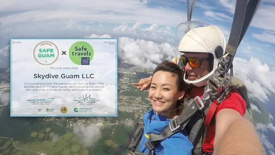 在關島高空跳傘的開心女體驗者與教練，後方是雲海與「Safe Guam」安全認證，確保跳傘安全可靠。