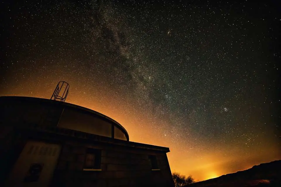 The Royal Observatory Greenwich at night, its iconic dome appearing mysterious in the faint light, set against a backdrop of a vast starry sky and city lights, blending scientific wonder with natural beauty.