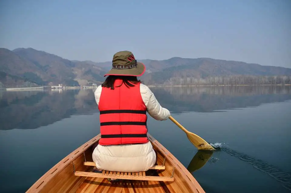 A solo visitor's back view while canoeing on Chuncheon Jungdo's tranquil lake, enjoying calm reflections and distant mountains, experiencing healing in nature.