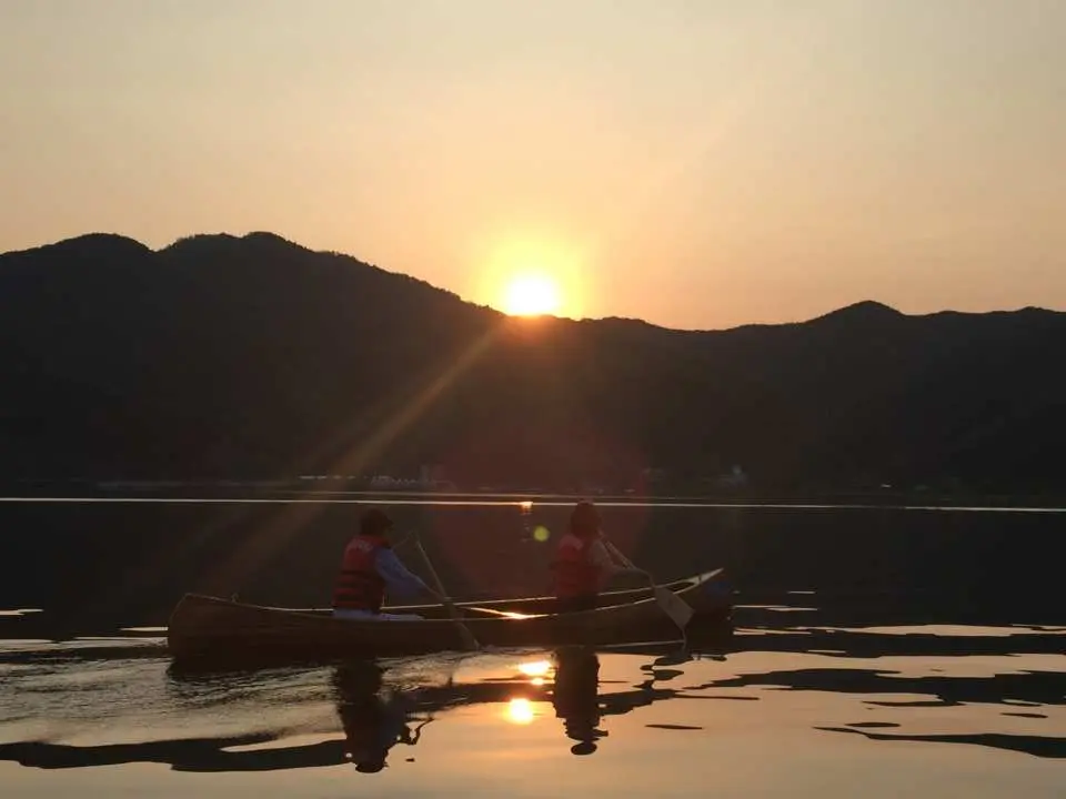 Two visitors on a Chuncheon Jungdo lake canoe watch the sunset, golden light shimmering on the water, creating a tranquil and romantic water experience near Seoul.