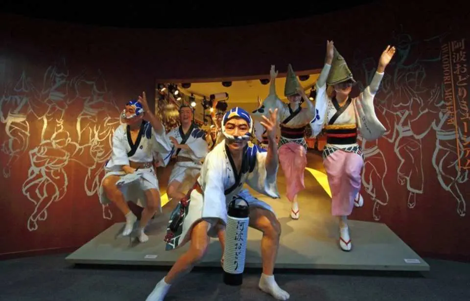 徳島阿波踊り会館で、来場者やパフォーマーが阿波踊りの伝統衣装を身につけ、歴史的な場面が描かれた壁画の前で、独特のダンス文化と情熱を体験。没入型のインタラクションを楽しんでいる。