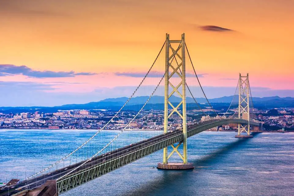 夕焼けに染まる日本の大鳴門橋。橋は温かい色彩に染まり、背景には美しい夕焼けと遠くの街の灯りが広がり、壮大で魅力的な景色は夕日鑑賞に最適。