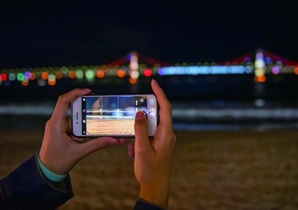 遊客在釜山海灘上用手機拍攝廣安大橋的璀璨夜景，橋身燈光絢麗，倒映在水面上，捕捉迷人海岸風光。