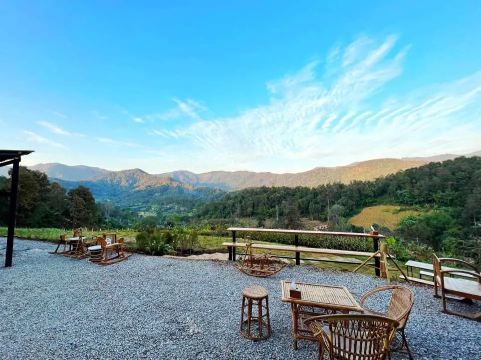 An outdoor cafe seating area at Elfin Farm Chiang Mai, with wooden tables and chairs facing majestic mountain views and a clear blue sky, perfect for relaxation.
